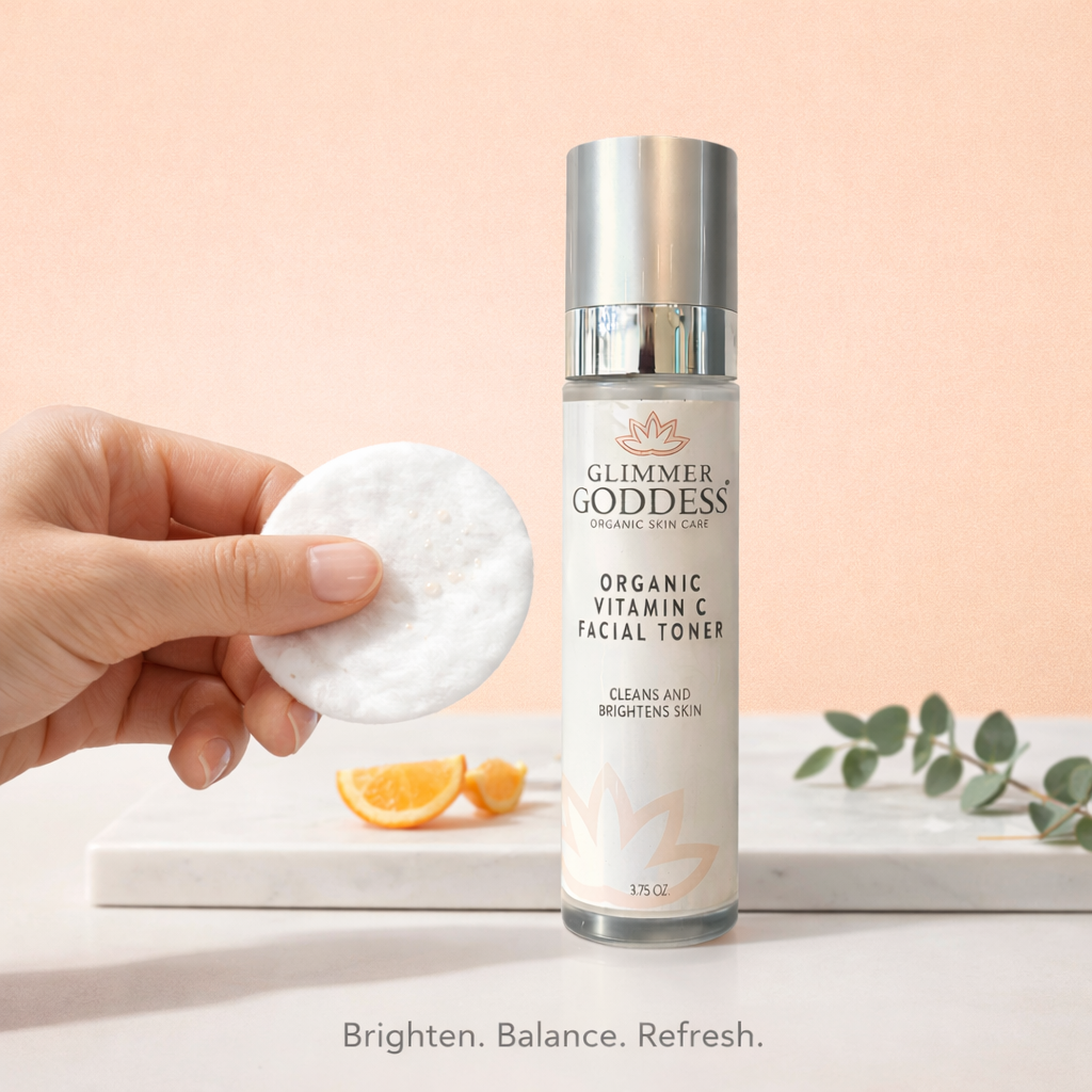 Glimmer Goddess® Organic Vitamin C Facial Toner (3.75 oz) in a luxury skincare vanity scene on a solid peach background, shown with a cotton round in use to brighten and even tone, refresh and balance skin, and help minimize the look of pores.