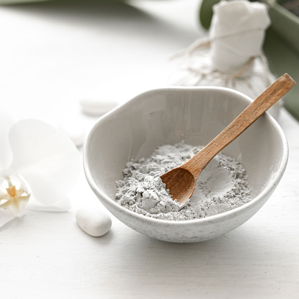 Bentonite clay powder in a white bowl with a wooden spoon, spa skincare ingredient